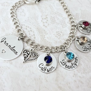May include: A silver charm bracelet with a heart charm and five round charms. Each charm has a different coloured gemstone and a name engraved on it. The names are Grandma, Bryson, Braelynn, Cayson, and Evelyn.