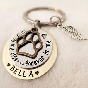 May include: A silver and gold keychain with a paw print charm, a small angel wing charm, and the text "Once my paw prints side...forever in my heart" and "BELLA".