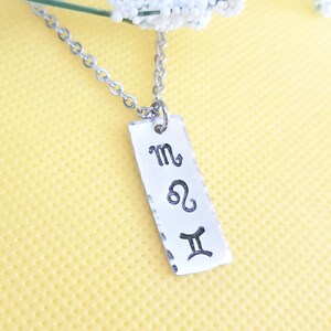 May include: Silver necklace with a rectangular pendant featuring the zodiac signs for Scorpio, Leo, and Gemini.