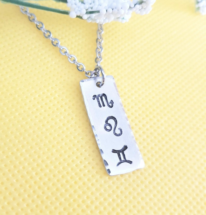May include: Silver necklace with a rectangular pendant featuring the zodiac signs for Scorpio, Leo, and Gemini.