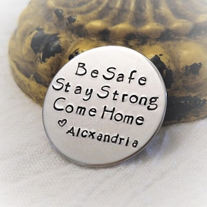 May include: A silver round charm with the text "Be Safe Stay Strong Come Home" and a heart symbol followed by the name "Alexandria".