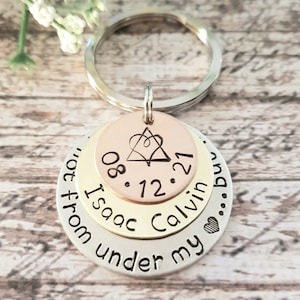 May include: A silver, gold, and rose gold layered keychain with the text "08.12.21 Isaac Calvin... from under my..." and a heart symbol.