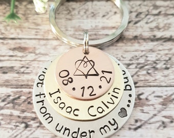 May include: A silver, gold, and rose gold layered keychain with the text "08.12.21 Isaac Calvin... from under my..." and a heart symbol.