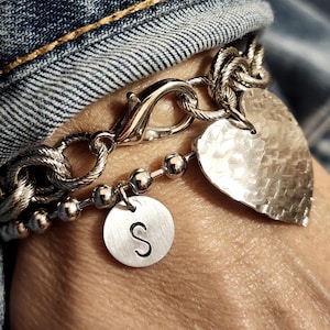 May include: Silver bracelet with a heart charm and a round charm with the letter "S". The bracelet has a textured heart-shaped charm, a chain link design, and a clasp. The bracelet is worn on a wrist.
