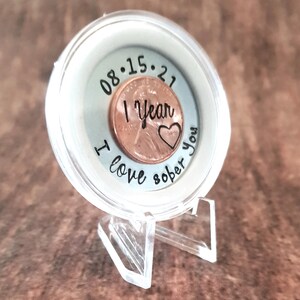 May include: A clear plastic coin display case with a copper penny inside. The penny has the date 08.15.21 engraved on it, along with the words "1 year sober" and a heart. The case has the words "I love sober you" engraved on the outside.