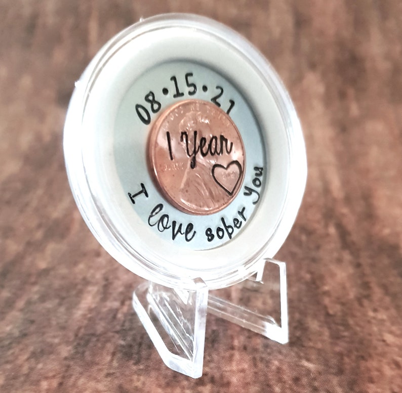 May include: A clear plastic coin display case with a copper penny inside. The penny has the date 08.15.21 engraved on it, along with the words "1 year sober" and a heart. The case has the words "I love sober you" engraved on the outside.