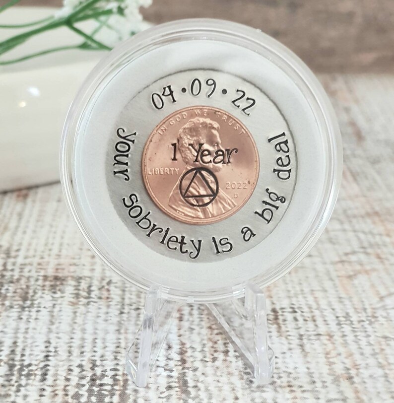 Op de afbeelding: Een herdenkingscent in een doorzichtige vitrine. De penny is gestempeld met "1 Year" en de datum "04.09.22". De tekst "Your sobriety is a big deal" is rond de penny gegraveerd. De behuizing staat op een transparante standaard.