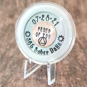 May include: A clear plastic coin holder with a copper penny inside. The penny has the words "Proud of You" and a triangle symbol engraved on it. The holder also has the date "07-28-21" and the text "365 Sober Days" around the edge.