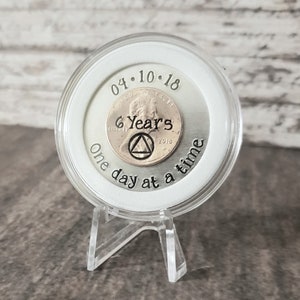 May include: A silver coin with the date 04.10.18 and the text "6 Years One day at a time" engraved on it. The coin is in a clear plastic display case with a white base.