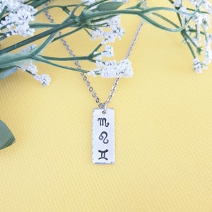 May include: A silver necklace with a rectangular pendant featuring the astrological symbols for Scorpio, Leo, and Gemini.