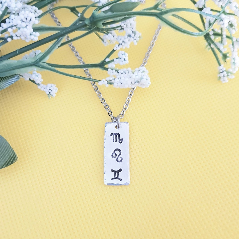May include: A silver necklace with a rectangular pendant featuring the astrological symbols for Scorpio, Leo, and Gemini.