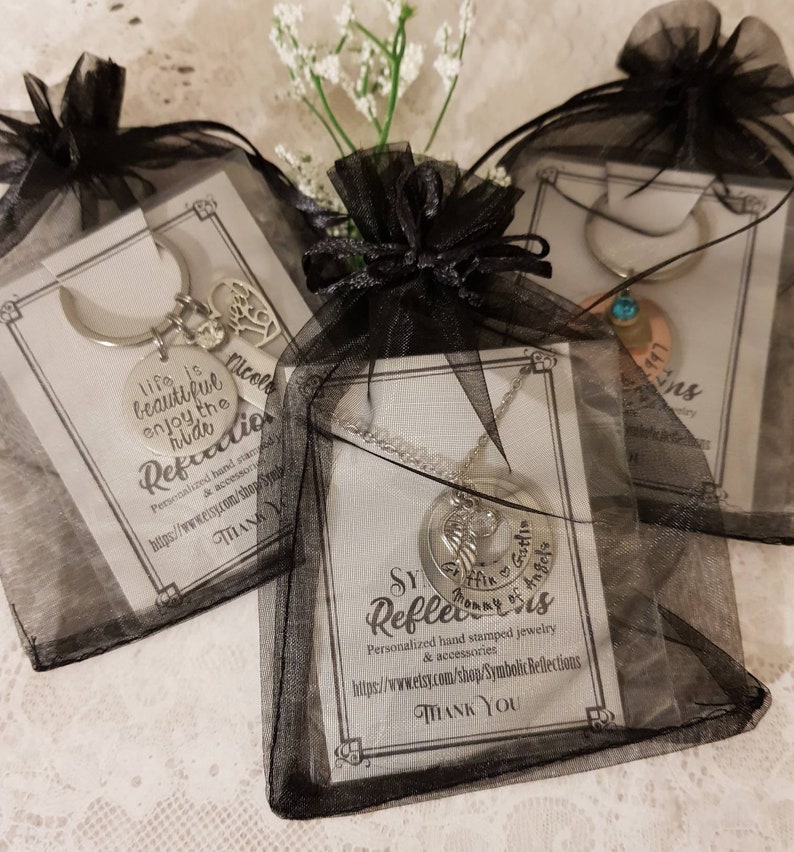 May include: Three black organza bags with silver jewelry inside. The bags are tied with black ribbon. The jewelry includes a necklace with a charm that says "Mommy of Angels", a keychain with a charm that says "Life is beautiful enjoy the ride", and a charm bracelet with a charm that says "Nicole".