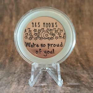 May include: A copper-colored coin with the text "365 Moons We're so proud of you!" surrounded by stars and a crescent moon. The coin is displayed on a clear plastic stand.