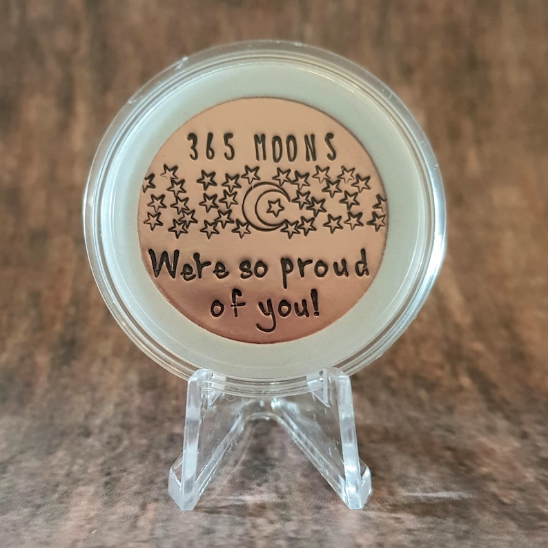 May include: A copper-colored coin with the text "365 Moons We're so proud of you!" surrounded by stars and a crescent moon. The coin is displayed on a clear plastic stand.