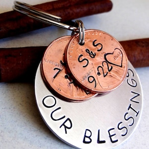 May include: A silver keychain with the words "Our Blessings" engraved on a round tag. Three copper pennies are attached to the tag, with the dates "9.22.2002" and "7.7" visible. The pennies also have the initials "S&S" engraved on them.