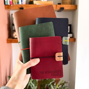 May include: A stack of four leather journals in tan, dark blue, forest green, and burgundy. The burgundy journal features a black elastic closure and the text THE HEMINGWAY'S TRAVELS embossed on the front cover. Each journal has a colored elastic band.