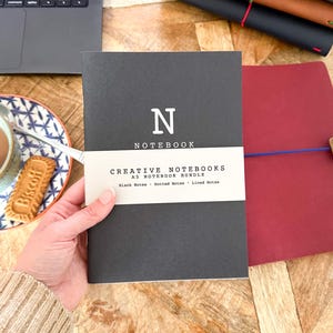 May include: A black A5 notebook with a white paper band featuring the text N NOTEBOOK, CREATIVE NOTEBOOKS, A5 NOTEBOOK BUNDLE, Blank Notes, Dotted Notes, Lined Notes. Additional notebooks in red, dark blue, brown, and green are visible nearby.
