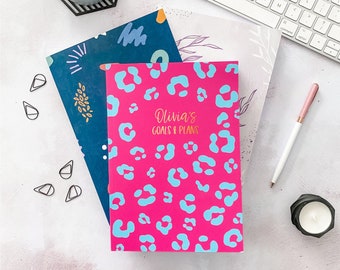 May include: A collection of notebooks with colorful covers, including a hot pink notebook with blue leopard print and the text "Olivia's Goals & Plans." Other notebooks feature abstract designs. A pen, candle, and keyboard are also visible.