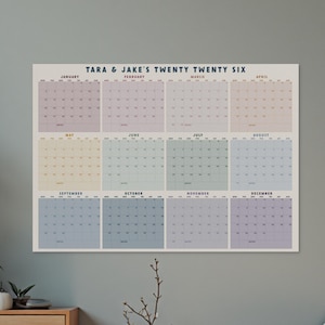 May include: A large wall calendar for 2026, with a pastel colour scheme for each month. The calendar is labelled "TARA & JAKE'S TWENTY TWENTY SIX" at the top. The calendar is rectangular and hangs on a grey wall.