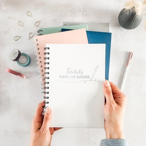 May include: A white spiral bound notebook with the text "Rachel's Plans for Success" on the cover. The notebook is being held by a person's hands.