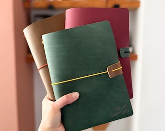 Op de afbeelding: Drie leren notitieboeken in groen, bruin en bordeauxrood. Het groene notitieboek heeft een gele elastische sluiting en de naam TAYLOR THOMPSON in de kaft gedrukt. Elk notitieboek is vastgezet met een gekleurde elastische band.