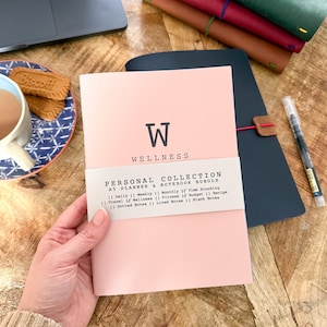 May include: A light pink A5 planner and notebook bundle with a white paper band featuring the text Personal Collection A5 Planner & Notebook Bundle and a checklist. The cover displays a large letter W and the word Wellness in black ink.