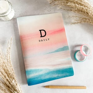 May include: A notebook with a watercolour design in pink, blue, and white. The cover displays the letter "D" and the word "DAILY" in black. Decorative washi tape and a pen are also visible.