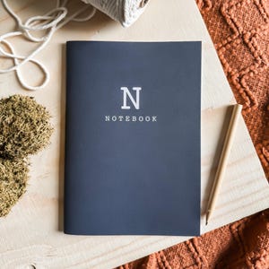 May include: A dark blue notebook with the letter "N" and the word "NOTEBOOK" printed in white on the front cover.