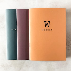 May include: Three notebooks with different colored covers. The notebook on the right is orange and has the text "W WEEKLY" on the front. The notebook in the middle is maroon and the notebook on the left is teal.