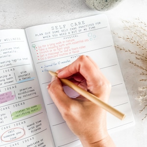 May include: An open planner with white pages featuring black printed text and lines. The page is titled SELF CARE and includes sections for physical and mental activities. A wooden pencil is held over the page, which contains handwritten text.
