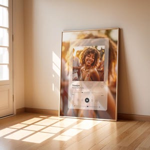 May include: A framed poster with a digital music player interface. The player is displaying the song "Titanium" by David Guetta ft. Sia. The background image is a blurry photo of a woman with curly hair smiling at a music festival.