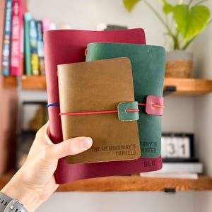 May include: Three stacked leather journals in burgundy, forest green, and tan. The tan journal is embossed with THE HEMINGWAY'S TRAVELS, the green with TONY'S NOTES, and the burgundy with BLH. Each features a colored elastic band closure.