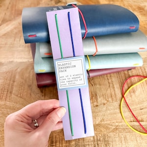 May include: An assortment of leather folios in various colors, stacked on a wooden surface. A package of elastic expansion ties in green and blue is in the foreground. The package reads "ELASTIC EXPANSION PACK".