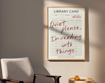 May include: A framed library card print with the text "Quiet please, I'm reading into things." The print has a beige background with a grid pattern and is in a light wood frame. A library card number is visible.