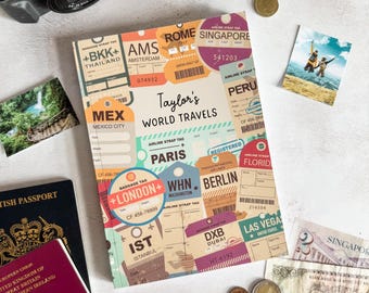 May include: A rectangular notebook titled Taylor's WORLD TRAVELS, featuring a collage of colorful airline baggage tags from cities like Bangkok, Rome, Singapore, Mexico City, Paris, London, Berlin, and Las Vegas, with various flight details.