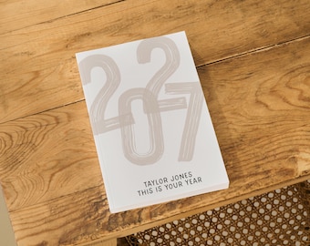 May include: A white rectangular planner with a soft beige brushstroke design featuring the year 2027 in large stylized numerals. The text Taylor Jones This Is Your Year is printed in black sans-serif font at the bottom of the front cover.