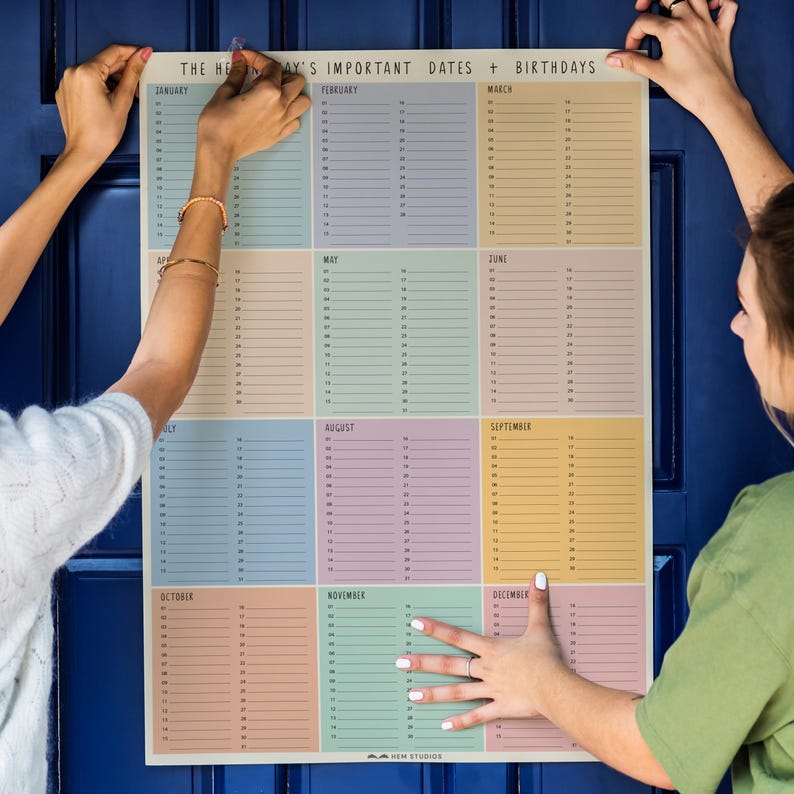 May include: A large, rectangular calendar with pastel-colored monthly sections. The calendar is titled "THE HEAY'S IMPORTANT DATES + BIRTHDAYS" and is being held up by two people. Each month has numbered lines for recording dates.
