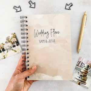 May include: A beige spiral bound notebook with the text "Wedding Plans" and "Sam & Jesse" on the cover. The notebook is held in a hand against a white background.