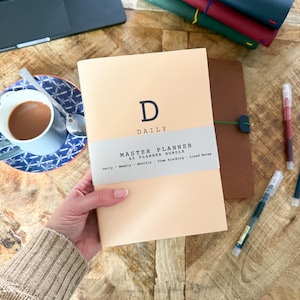 May include: A light peach-coloured A5 daily master planner with a grey paper band featuring the text D DAILY MASTER PLANNER A5 PLANNER BUNDLE Daily Weekly Monthly Time Blocking Lined Notes. The planner is held in a hand against a wooden surface.