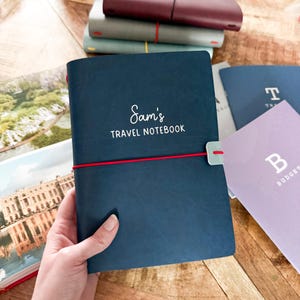 May include: A navy blue travel notebook with the text "Sam's TRAVEL NOTEBOOK" in white script. A red elastic band secures the notebook, and a small green clasp is visible. Other notebooks in various colors are stacked in the background.