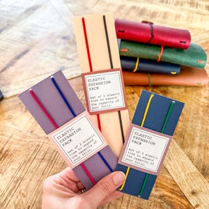 May include: Three rectangular cardstock holders each containing two elastic bands. The cards are light purple, beige, and dark blue, labelled ELASTIC EXPANSION PACK with text describing the set of 2 elastic ties to expand the capacity of your folio.
