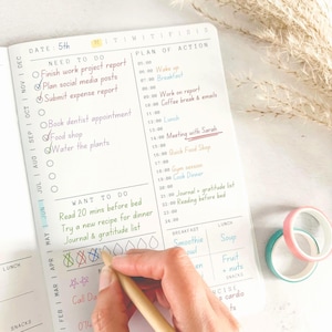 May include: A daily planner page with sections for Need To Do, Plan Of Action, and Want To Do. The page features handwritten tasks in various colored inks, including checkmarks and crossed-out icons. Two rolls of washi tape in pink and teal sit nearby.