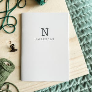 May include: A white notebook with a dark gray letter N printed at the top center and the word NOTEBOOK printed in smaller dark gray capital letters directly below it. The notebook has a clean, simple cover design with a vertical spine on the left.