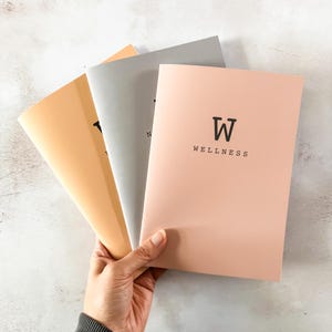 May include: Three notebooks with different coloured covers. The covers are peach, grey, and light brown. The peach notebook has the word "WELLNESS" and a capital "W" printed on the front.