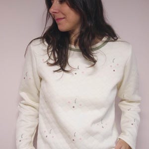 May include: Cream-colored quilted sweatshirt with long sleeves and a crew neck. The sweatshirt features a subtle floral pattern in pink and brown. The neckline has a green trim. The person is wearing blue jeans.