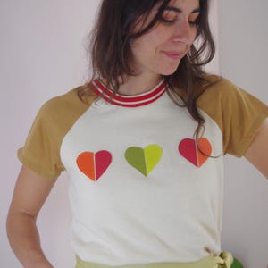 Peut inclure: T-shirt blanc avec des manches moutarde et un col rayé rouge et blanc. Le t-shirt présente trois appliqués en forme de cœur colorés. Le modèle porte une jupe vert clair.