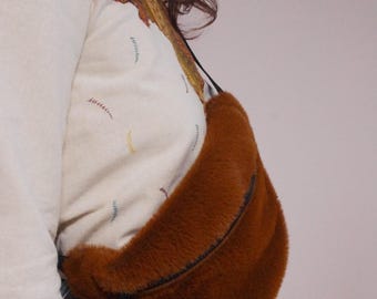 May include: A brown faux fur fanny pack with a black zipper and strap. The fanny pack is worn around the waist of a person wearing a white shirt and blue jeans.