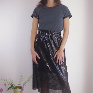 Peut inclure: Une personne porte un t-shirt gris foncé et une jupe à sequins noire. La jupe arrive au genou et scintille. Des bottines noires complètent la tenue. Un trophée avec des fleurs est en arrière-plan.