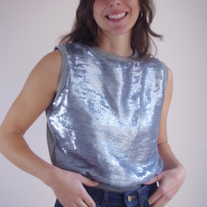 Può includere: Un top senza maniche con paillettes argentate e rifiniture grigie. Il top è abbinato a jeans di denim blu scuro. La modella sorride, con le mani nelle tasche dei jeans. Il top è un capo scintillante e accattivante.