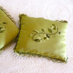 May include: Two square, lime green cushions with embroidered floral designs. The cushions have a scalloped trim in a darker green and gold. The cushions are resting on a white, textured surface.